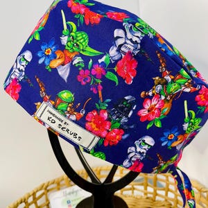 May include: A navy blue scrub cap with a colourful pattern of Star Wars characters and flowers. The cap features Yoda, Stormtroopers, and Boba Fett amongst red, pink, and blue floral designs. The label reads "Handmade by KD Scrubs".