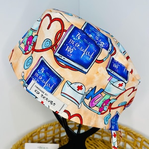 May include: A colourful surgical scrub hat with a light orange background. The hat features a repeating pattern of hearts, stethoscopes, and medical monitors with the word "Nurse" in a colourful font. The hat has ties and a label that reads "Handmade by: KD Scrubs".