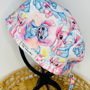 May include: A white scrub hat with a colourful watercolour background featuring cartoon Stitch characters. The hat has a tie at the back and says "Handmade by: KD Scrubs" on the side.
