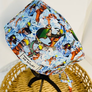 May include: A light blue scrub cap with a repeating pattern of orange and black anime characters. The cap has a white tag that reads "HANDMADE BY: KD SCRUBS". The cap is tied at the back. The cap is made of fabric.