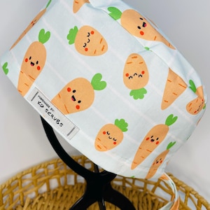 May include: A white scrub hat with a light blue and white striped pattern and a repeating pattern of orange carrots with green tops and happy faces. The hat has a tie and a label that reads "Handmade by KD Scrubs".