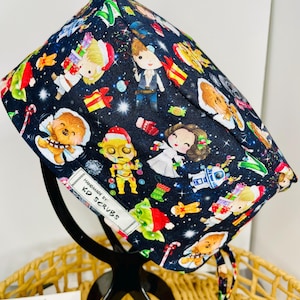 May include: A navy blue scrub cap with a Star Wars-themed festive print. The design includes characters in holiday attire, such as Santa Claus and Christmas presents. The cap has a white tag that reads "HANDMADE BY KD SCRUBS".