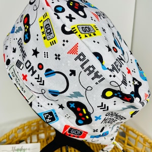 May include: White scrub cap patterned with video game controllers and phrases. The design includes the words "Game On", "Play", and "GO!". The cap also features headphones and other gaming-related graphics.