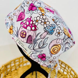 May include: White surgical scrub hat with a whimsical mushroom and snail print. The hat has pink, blue, and yellow accents. The hat has a label that says "Handmade by KD Scrubs".