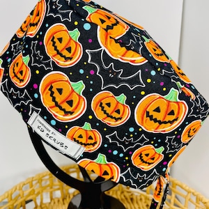 May include: A black Halloween scrub cap with a repeating pattern of orange pumpkins, white bats, and colourful polka dots. The cap has a white tag that reads "HANDMADE BY KD SCRUBS". The cap is displayed on a black stand.