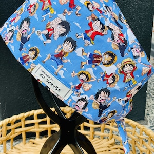 May include: A blue scrub cap with a repeating cartoon character design in action poses against a sky background. The cap has a white tag that reads "HANDMADE BY: KD SCRUBS".