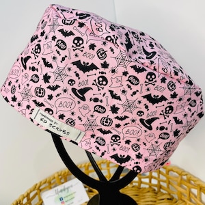 May include: A pink Halloween scrub cap featuring a black pattern of bats, spiderwebs, skulls, ghosts, and the word "BOO!". The cap has a white tag that reads "HANDMADE BY: KD SCRUBS".