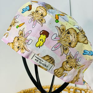May include: A scrub cap with a repeating pattern of cartoon bunnies, carrots, and Easter-themed designs. The cap is in pastel pink and yellow with white stripes. The label reads "HANDMADE BY: KD SCRUBS."