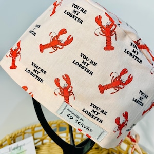 May include: A light pink scrub cap featuring a pattern of red lobsters and the words "YOU'RE MY LOBSTER". The cap includes a tag that reads "HANDMADE BY: KD SCRUBS".