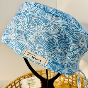 May include: A blue and white patterned scrub cap with a wave design. The cap has a tag that reads "HANDMADE BY: KD SCRUBS". The cap is displayed on a black stand.