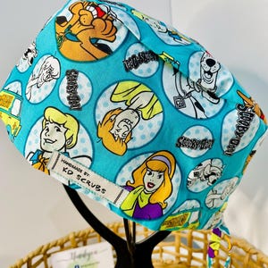 May include: A turquoise scrub cap with cartoon characters from Scooby-Doo in white polka dots. The cap has a tag that reads "HANDMADE BY: KD SCRUBS". The cap is tied with a colourful ribbon.