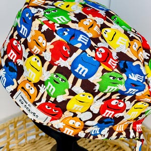 May include: A brown scrub hat with a colourful M&M's pattern. The hat has a tie on the back and a white label that says "Handmade by KD Scrubs".