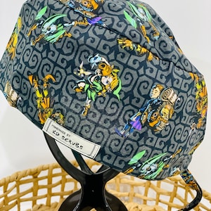 May include: A dark grey scrub cap with a repeating pattern of video game characters in yellow, green, and purple. The cap has a white tag that reads "HANDMADE BY: KD SCRUBS". The cap is displayed on a black stand.