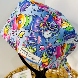 May include: A light blue scrub cap with a vibrant cartoon character design and the word "LOVE" in bubble letters. The cap features various cartoon characters and the text "HANDMADE BY: KD SCRUBS".
