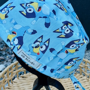 May include: Blue surgical scrub hat featuring a repeating pattern of a cartoon blue dog with yellow ears and a red tongue. The hat has a white label that reads "Handmade by KD Scrubs".