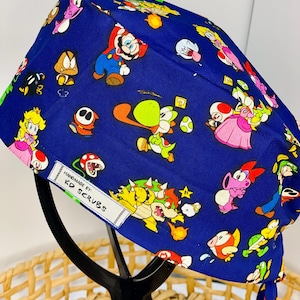 May include: Blue surgical scrub hat with a colourful Super Mario Bros. print. The hat has ties and a label that reads "Handmade by KD Scrubs".