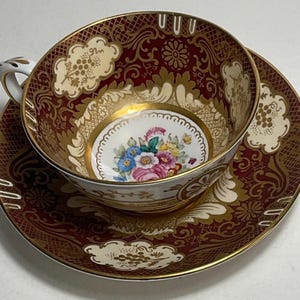 May include: A porcelain teacup and saucer set featuring a deep burgundy background with intricate gold filigree patterns. The interior of the cup and the center of the saucer display a colorful floral bouquet on a white base with gold accents.