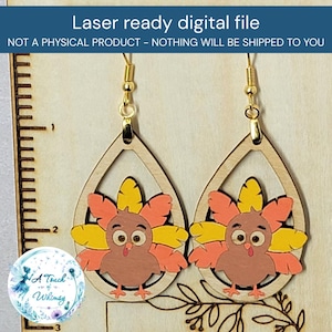 May include: A pair of teardrop-shaped wooden earrings featuring a cartoon turkey with yellow, orange, and brown feathers. The earrings hang from gold-toned metal hooks and are positioned next to a ruler showing a measurement of 3 inches.
