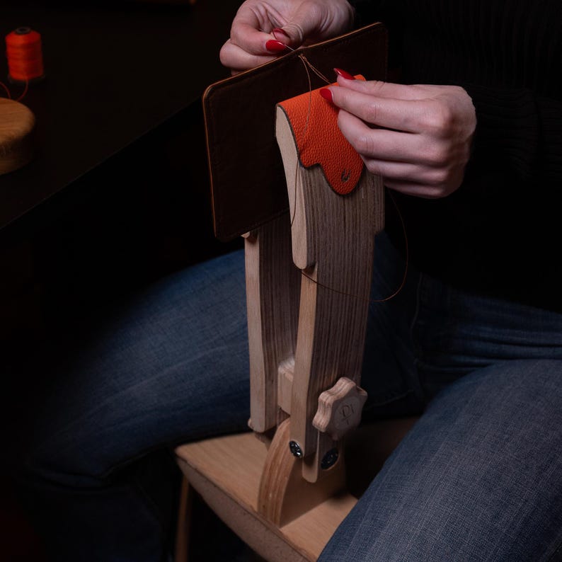 May include: A wooden leather stitching pony holding a dark brown leather piece with an orange leather accent. The tool features a light wood grain finish, a central tension adjustment knob, and a base that rests on a seat for stability during use.
