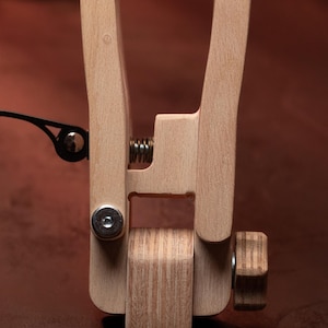 May include: A light-colored wooden clamp mechanism featuring a central metal spring, a silver-toned circular bolt, and a textured wooden knob on the side. The structure consists of two vertical wooden arms connected by a pivot point and a tension spring.