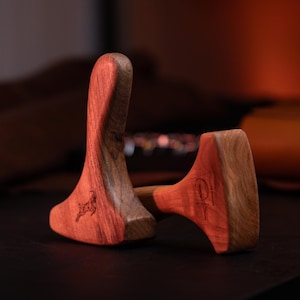 Könnte beinhalten: Zwei glatte, rotbraune Massagewerkzeuge aus Holz mit geschwungener, ergonomischer Form. Ein Werkzeug zeigt eine kleine, dunkle Gravur einer Ziege, während das andere ein kreisförmiges Logo mit dem Schriftzug "JASMINA" und einem Symbol trägt.