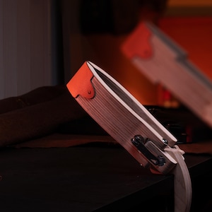 May include: A wooden stitching pony for leatherworking featuring a light wood grain finish, a bright orange textured leather top grip, and a black metal tension adjustment screw with a wing nut mechanism for securing leather pieces during hand stitching.