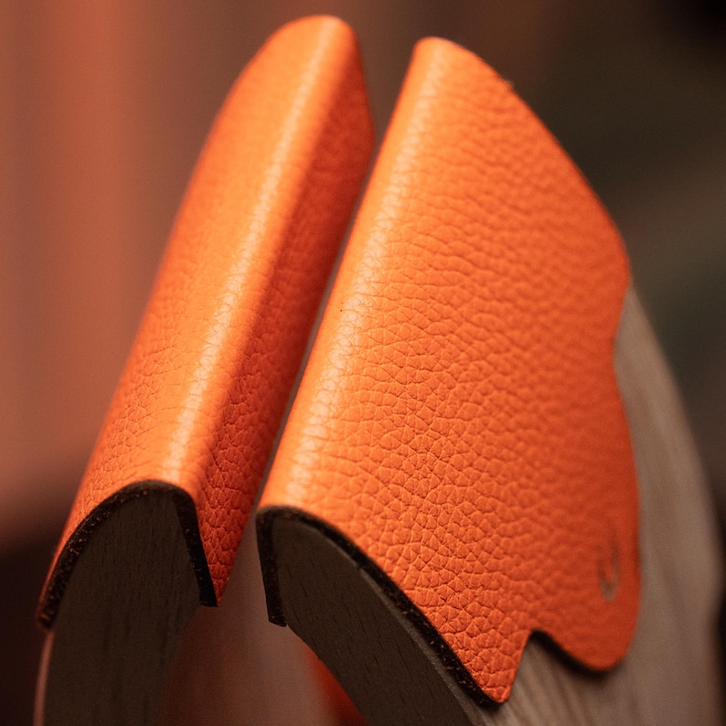May include: Two curved wooden pieces featuring bright orange textured leather overlays on their outer edges. The leather has a distinct pebbled grain and is securely attached to the dark wood, showing a clean edge and a smooth, rounded profile.