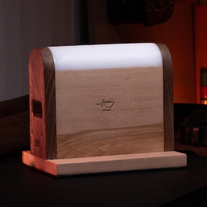 Peut inclure: Une lampe rectangulaire en bois composée d'un panneau central en bois clair, d'un cadre en bois foncé et d'une section supérieure blanche translucide. Le panneau central présente un logo noir avec le texte "H" et "DESIGN CO." en petit.