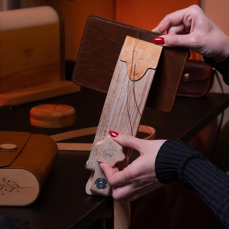 May include: A wooden leather stitching pony featuring a light-colored wood frame, a central tensioning knob with a star shape, and a leather-lined clamp. The tool holds a piece of dark brown leather with a visible thread for hand-stitching projects.