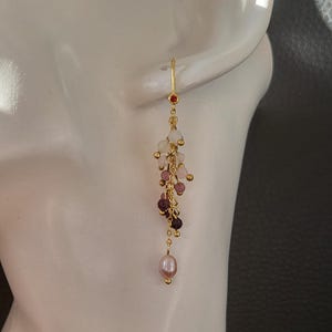 Könnte beinhalten: Goldene Ohrhänger mit einer rosafarbenen Perle am unteren Ende. Die Ohrringe haben eine Kaskade aus kleinen, facettierten Edelsteinen in Weiß-, Rosa- und Lilatönen.