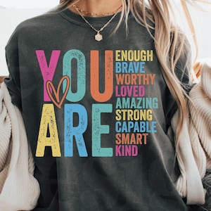 May include: A gray sweatshirt with the words "YOU ARE" in colorful letters. The words "enough, brave, worthy, loved, amazing, strong, capable, smart, kind" are stacked vertically to the right of the words "YOU ARE".