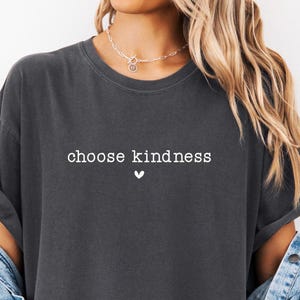 May include: Dark gray t-shirt with the words "choose kindness" in white, accompanied by a small white heart. The shirt has a relaxed fit and is paired with a light blue denim jacket.