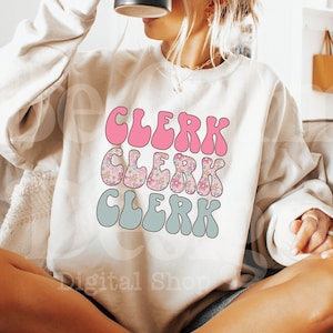 Può includere: Una felpa bianca con il testo "CLERK" ripetuto tre volte in un font retrò e colorato. Il testo è in rosa, blu e verde con un motivo floreale.