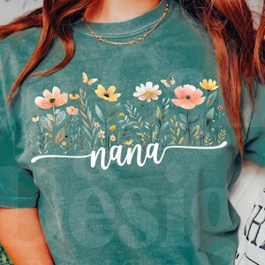 Pode incluir: Uma camiseta verde com um design floral e a palavra "nana" impressa nela. A camiseta está sendo usada por uma pessoa sentada em um sofá.