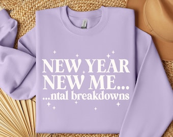May include: A lilac sweatshirt featuring the white text "NEW YEAR NEW ME... mental breakdowns". The garment is placed on a woven mat, with a brown hat visible. The design includes star accents.