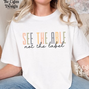 May include: White t-shirt with the text "SEE THE ABLE not the label" in colorful letters. The text is in a vertical stack with the word "ABLE" in gray, "SEE" in green, "THE" in orange, and "not the label" in black.
