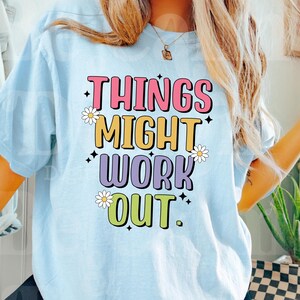 Puede incluir: Una camiseta azul claro con el texto "Things Might Work Out" en letras de colores con margaritas blancas alrededor del texto.