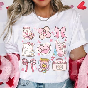 Puede incluir: Camiseta blanca con un diseño rosa y blanco que presenta varios símbolos médicos, incluyendo un corazón, una venda, un frasco de pastillas, un estetoscopio y un botiquín de primeros auxilios. El diseño está rodeado de pequeños corazones y estrellas rosas.