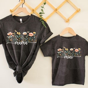 Puede incluir: Dos camisetas de color gris oscuro con un diseño floral y las palabras "mama" y "mini" impresas en ellas. Las camisetas están colgadas en un estante de madera.