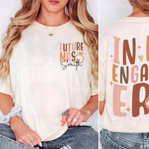May include: A white t-shirt with the text "Future Mrs. Smith" on the front and "In My Engaged Era" on the back. The text is in a brown, pink, and gold colour scheme with a diamond ring graphic.