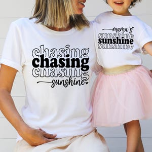 May include: Two white t-shirts with black text. The larger shirt says "chasing chasing chasing sunshine" and the smaller shirt says "mama's sunshine sunshine sunshine".