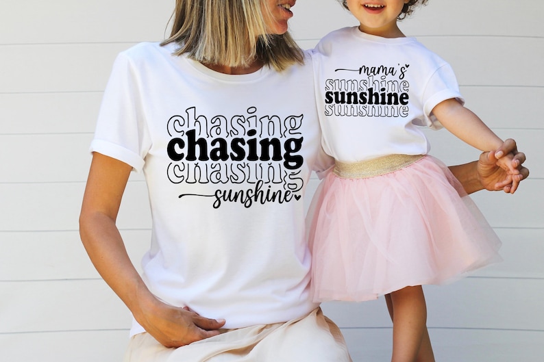 May include: Two white t-shirts with black text. The larger shirt says "chasing chasing chasing sunshine" and the smaller shirt says "mama's sunshine sunshine sunshine".