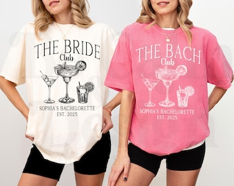 May include: Two pink and white t-shirts with a bachelorette party theme. The text on the shirts reads "The Bride Club" and "The Bach Club". Both shirts feature a graphic of three cocktails with olives and a lime.