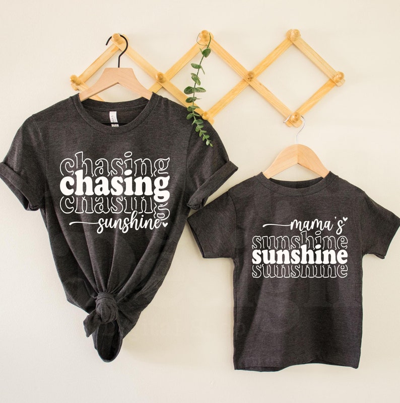 May include: Two gray t-shirts with white text. The larger shirt says "chasing chasing chasing sunshine" and the smaller shirt says "mama's sunshine sunshine sunshine".