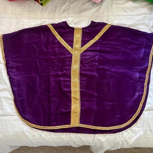 May include: A deep purple liturgical vestment with gold trim. The garment has a wide, cape-like design with a rounded hem. Gold trim accents the neckline, front, and edges, creating a striking contrast against the rich purple fabric.