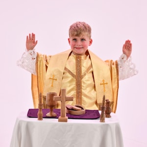 May include: A gold-colored liturgical vestment featuring white lace cuffs and embroidered cross motifs, worn with a matching stole. On the table are a wooden chalice, a wooden cross, and a small wooden bowl containing circular communion wafers.