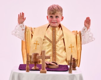 Może przedstawiać: Złota szata liturgiczna z białymi koronkowymi mankietami i haftowanymi motywami krzyża, noszona z pasującą stułą. Na stole znajduje się drewniany kielich, drewniany krzyż oraz mała drewniana miseczka z komunikantami w kształcie koła.