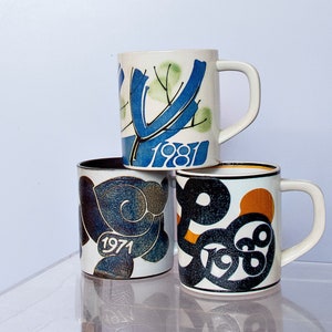 Può includere: Tre tazze in ceramica con diversi disegni. La tazza superiore ha un design astratto blu e verde con l'anno 1981. La tazza centrale ha un design astratto blu e marrone con l'anno 1971. La tazza inferiore ha un design astratto nero e arancione con l'anno 1980.
