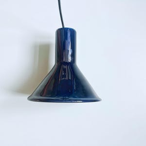 Puede incluir: Una lámpara colgante de color azul marino profundo y brillante con una pantalla cónica lisa y una sección superior cilíndrica. La lámpara está suspendida por un cable negro fino y presenta una superficie reflectante que capta la luz.