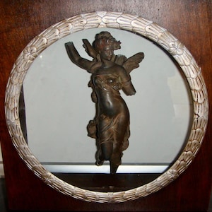Puede incluir: Una estatua de ángel en tono bronce, sin brazos, está enmarcada dentro de un borde circular plateado. El ángel está sobre un fondo transparente, encerrado en un marco de madera oscura. Una obra de arte clásica.